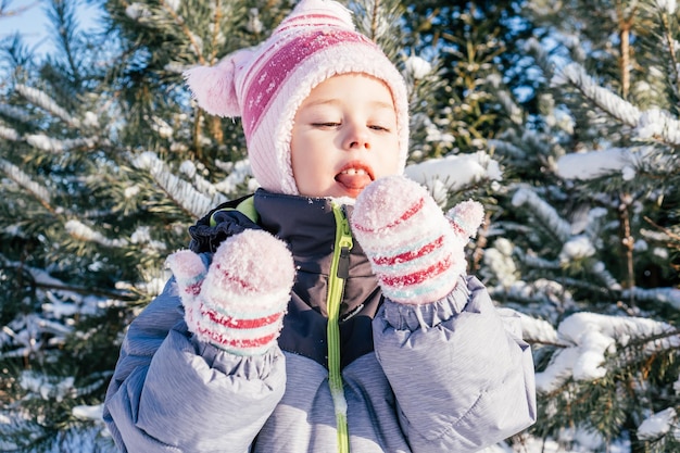 Little girl 34 years old in winter overalls hat and mittens stands against snowcovered pines and fir trees eats snow