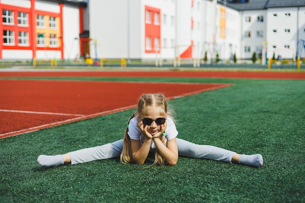 A little girl of 78 years old in a white Tshirt and sunglasses is sitting on the sports field The girl is sitting on the green grass on a sunny day