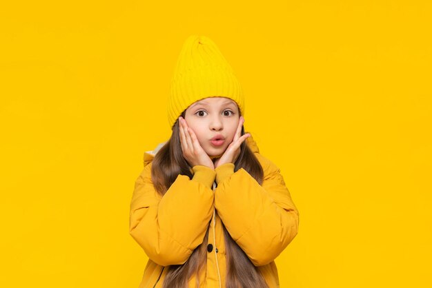 A little girl in a beanie hat and an orange jacket pursed her lips in surprise Warm autumn clothes for teenagers and children The child is surprised Yellow isolated background