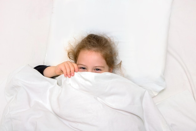 Little girl in bed is covered with a blanket only laughing eyes are visible