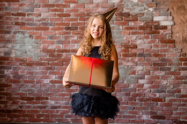 A little girl in a birthday cap holds a gold gift box and rejoices.