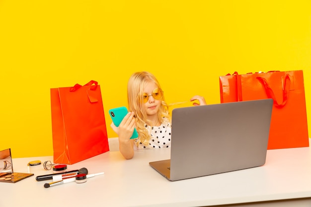 Little girl blogger with blue phone making video for blog and followers sitting in the yellow studio isolated.