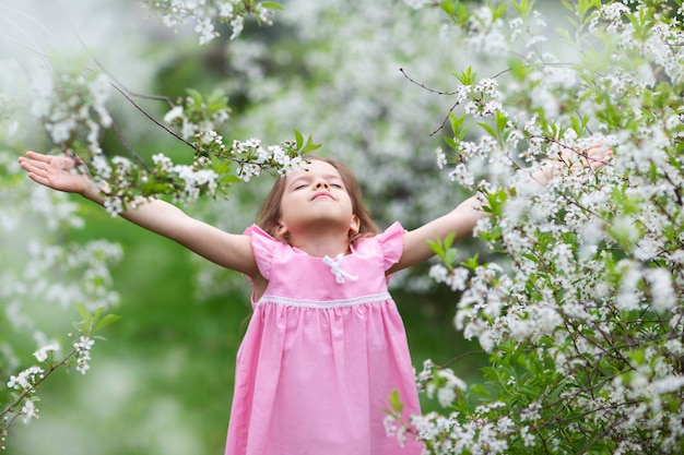 A little girl in a blooming garden, closes her eyes and stretches her hands to the sky