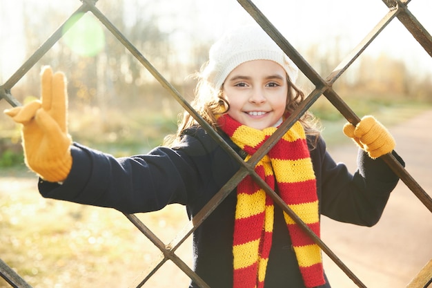 Little girl in a blue coat behind a metal lattice in the park in autumn. High quality photo