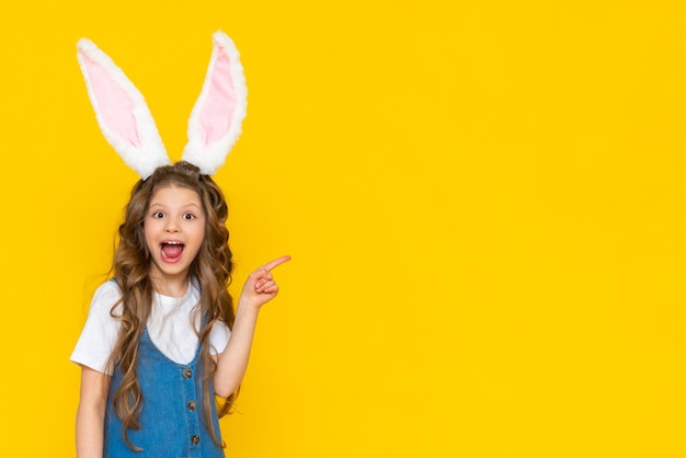 A little girl in a blue dress with bunny ears points to the side on a yellow background.