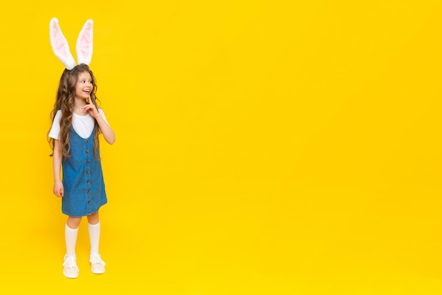 A little girl in a blue dress with a rabbit ears stands on a yellow background.