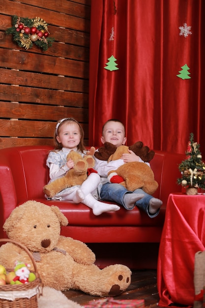 Little girl and boy on sofa in christmas studio in red colors. christmas tree and teddy bear on wall.