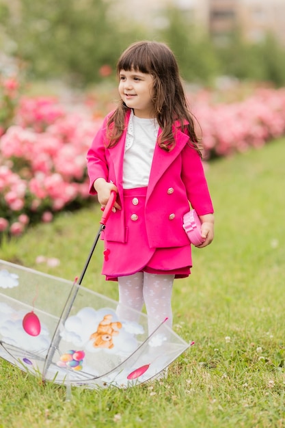 Little girl in a bright fuchsia suit walks outside under a transparent umbrella card banner