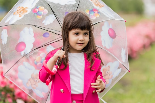 Little girl in a bright fuchsia suit walks outside under a transparent umbrella card banner