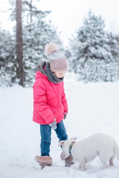 Little girl in a bright jacket plays in the winter snowy forest with her dog jack russell terrier