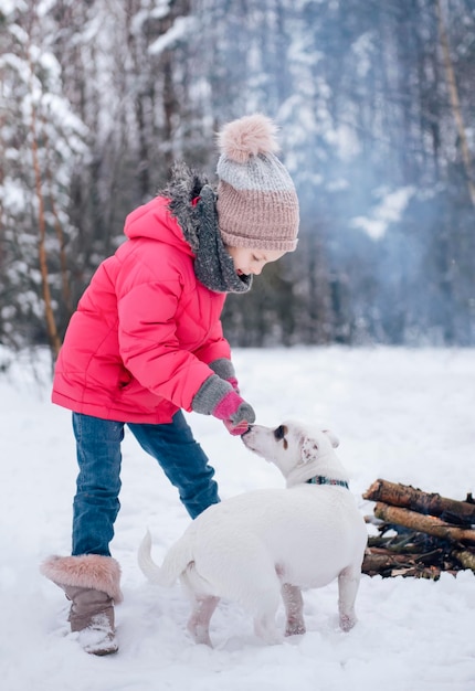 Little girl in a bright jacket plays in the winter snowy forest with her dog jack russell terrier