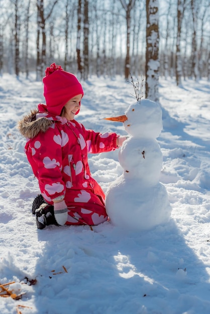 Little girl building snowman in snowy park Active outdoors leisure with family with children in winter Kid during stroll in a snowy winter park