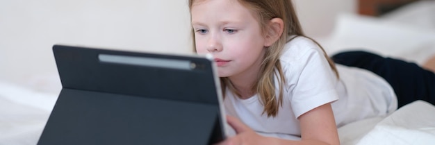 Little girl carefully looks at tablet while lying on bed