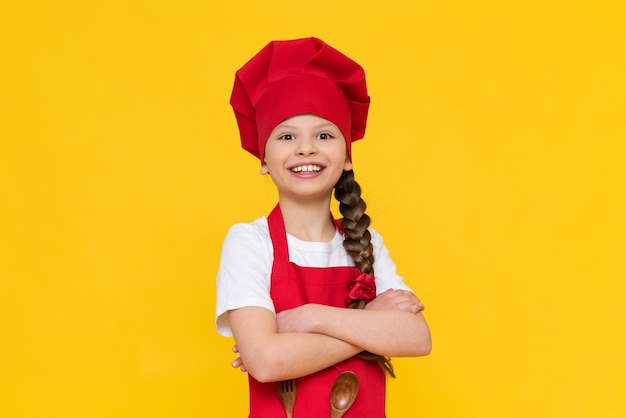 A little girl in a chef costume is learning to cook Cooking by children A child in a red apron and a cap on a yellow isolated background