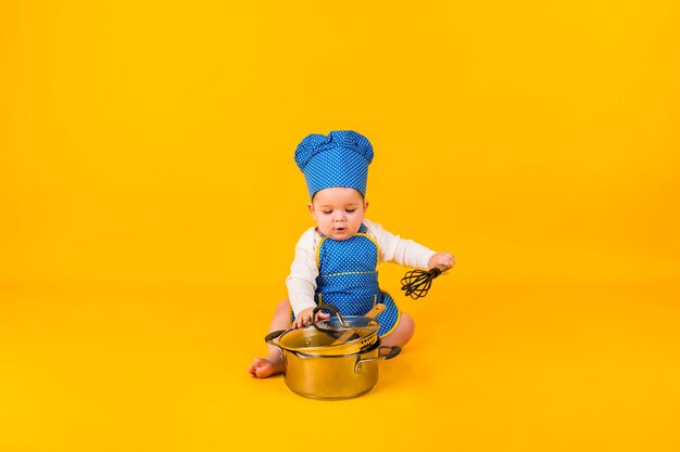 A little girl in a chef costume sits with a metal pot on a yellow wall with space for text