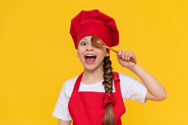 A little girl chef is playing with wooden spoons Cooking for children A child in a red apron and a cap on a yellow isolated background