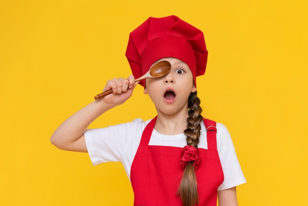 A little girl chef is playing with wooden spoons Cooking for children A child in a red apron and a cap on a yellow isolated background