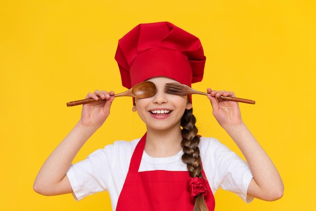 A little girl chef is playing with wooden spoons Cooking for children A child in a red apron and a cap on a yellow isolated background