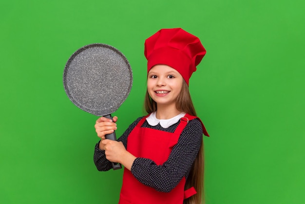 A little girl chef in a red hat and apron holds a pancake pan on a green isolated background Choosing a profession for a student