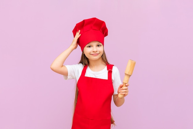 A little girl in a chef's hat is preparing for baking holding a rolling pin in her hands to roll out the dough Baby baker on a pink isolated background