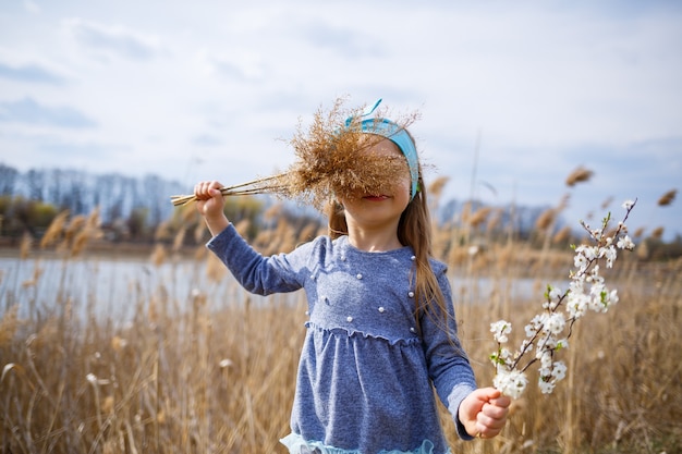 Little girl child holds dry reeds and a branch with small white flowers in hands, sunny spring weather, smilling and joy of the child