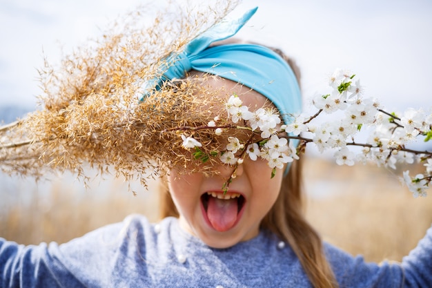 Little girl child holds dry reeds and a branch with small white flowers in hands, sunny spring weather, smilling and joy of the child