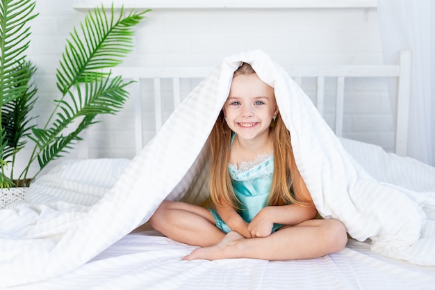 A little girl child is playing under the blanket waking up in the morning at home on the bed on a white cotton bed and smiling sweetly