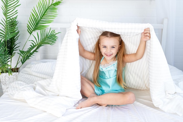 A little girl child is playing under the blanket waking up in the morning at home on the bed on a white cotton bed and smiling sweetly