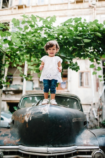 The little girl child is standing on ancient vintage car Young happy family during a walk the street of old city town outdoors The concept of family holiday and travel The mom holds little girl