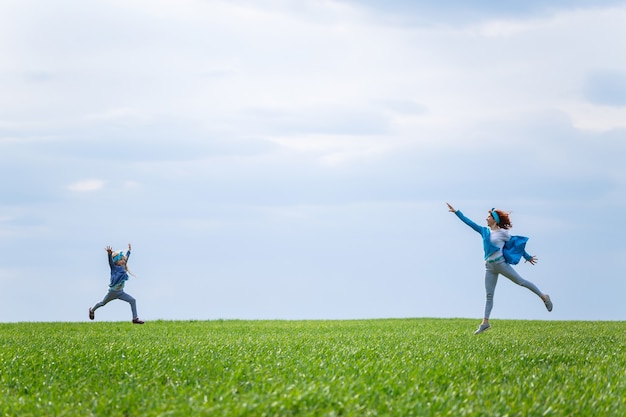 Little girl child and mother woman run and jump, green grass in the field, sunny spring weather, smile and joy of the child, blue sky with clouds