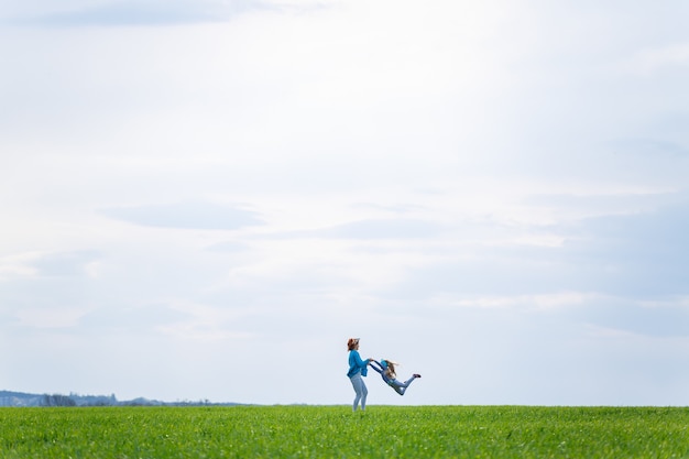 Little girl child and mother woman run and jump, green grass in the field, sunny spring weather, smile and joy of the child, blue sky with clouds