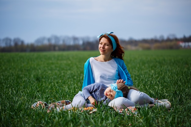 Little girl child and mother woman sit on the bedspread, green grass in the field, sunny spring weather, smile and joy of the child, blue sky with clouds