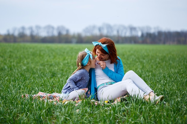 Little girl child and mother woman sitting on the bedspread and eating cookies and marmalade, green grass in the field, sunny spring weather, smile and joy of the child, blue sky with clouds