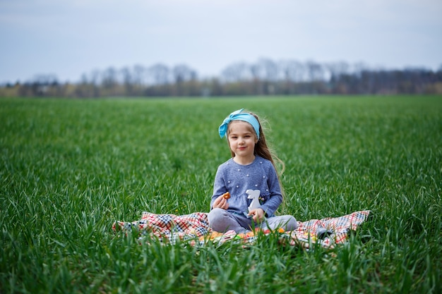 Little girl child sits on the bedspread and eats cookies and marmalade, green grass in the field, sunny spring weather, smile and joy of the child, blue sky with clouds