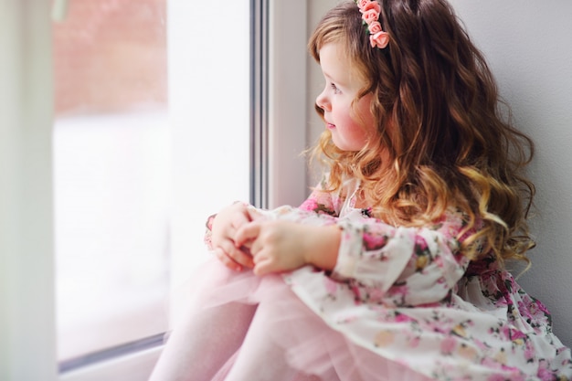 little girl the child in a smart dress sits on the windowsill, looks out the window and is sad.