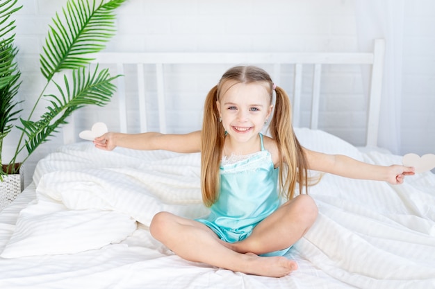 Little girl child with hearts in her hands sitting at home on the bed on a white cotton bed and smiling sweetly, valentine's day concept