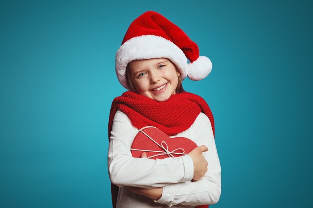 Little girl in christmas hat red scarf embracing present box in shape of