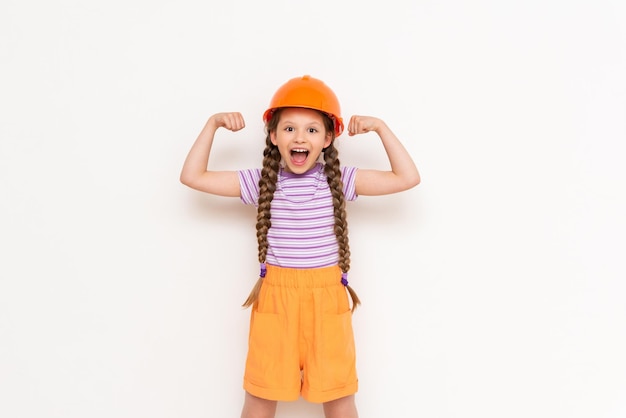 A little girl in a construction helmet and pigtails shows the strength of her biceps on a white isolated background The profession of an engineer for a child