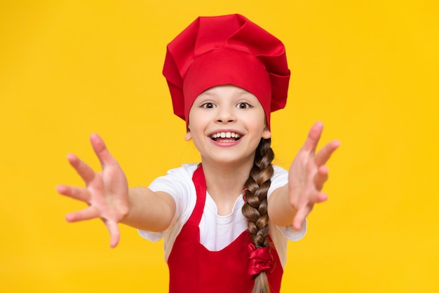 The little girl cook stretches her arms forward and smiles the concept of a child preparing food at home a beautiful attractive girl on a yellow isolated background in the cook's costume