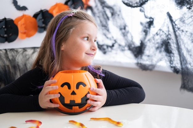 little girl in costume of witch holding pumpkin jack with candies, celebrating Halloween at home