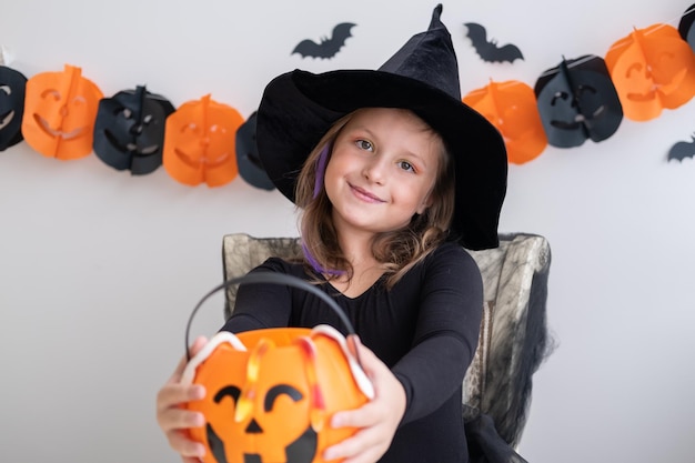 little girl in costume of witch holding pumpkin jack with candies, celebrating Halloween at home