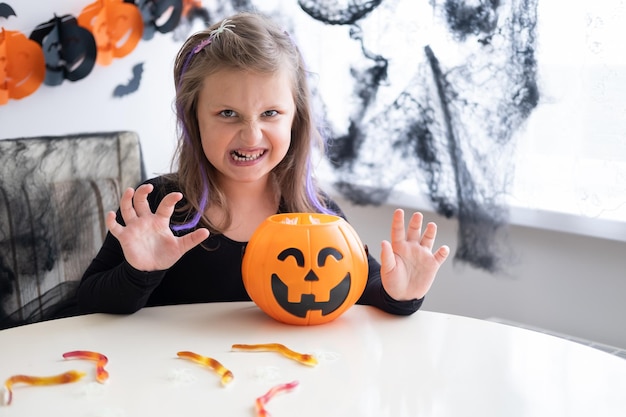 little girl in costume of witch holding pumpkin jack with candies, celebrating Halloween at home