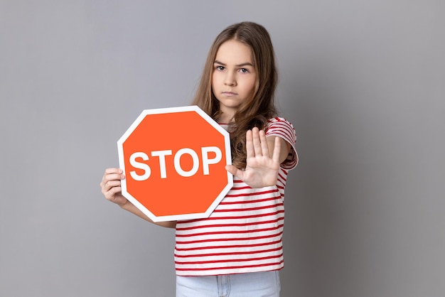 Little girl covering face with octagonal Stop symbol showing traffic sign and ban gesture with palm