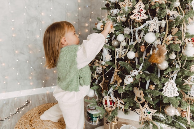 Little girl decorates the Christmas tree with toys. A cute baby is preparing at home for the celebration of Christmas.