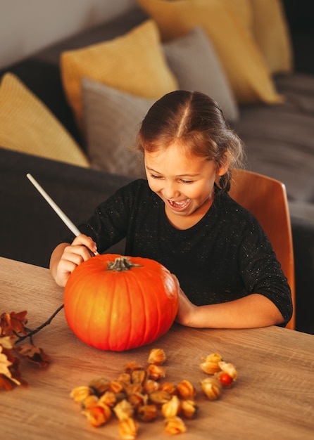 Photo a little girl decorates a pumpkin in a room for halloween. kids decorate home. children carve pumpkin. spooky halloween fun