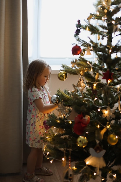 Little girl decorating christmas tree with toys and baubles Cute kid preparing home for xmas celebration