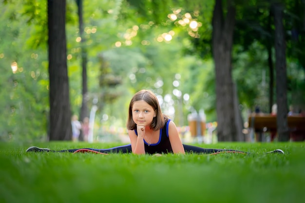 Little girl doing yoga sits on the lawn in the park in a twine and looks at the camera