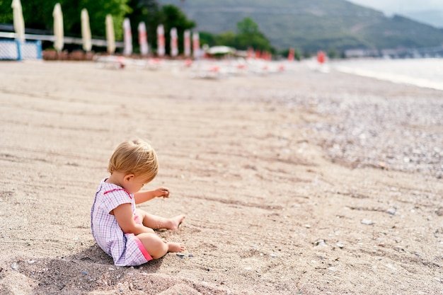 Little girl in a dress plays with sand on the beach side view