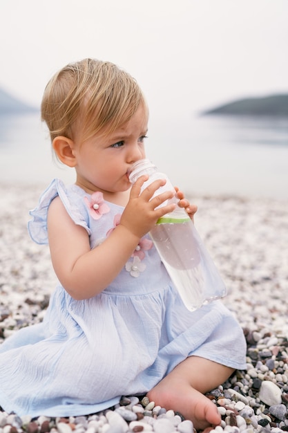 Little girl in a dress sits on a pebble beach and drinks water from a bottle