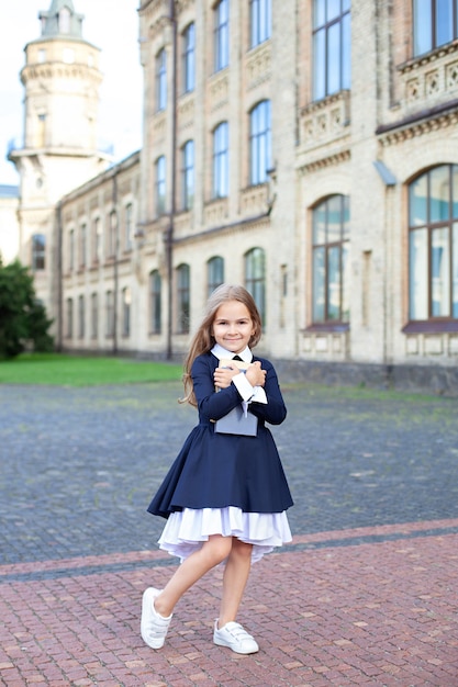 Little girl in dress and with long hair outdoors near school building. Child with books.